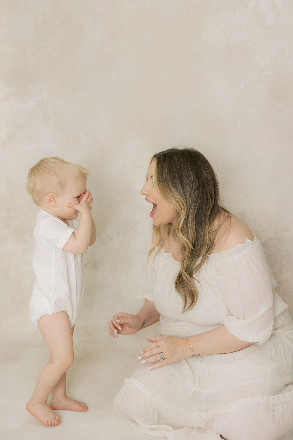 A toddler boy in a white onesie plays peek-a-boo with mom while she sits in a studio in a white dress
