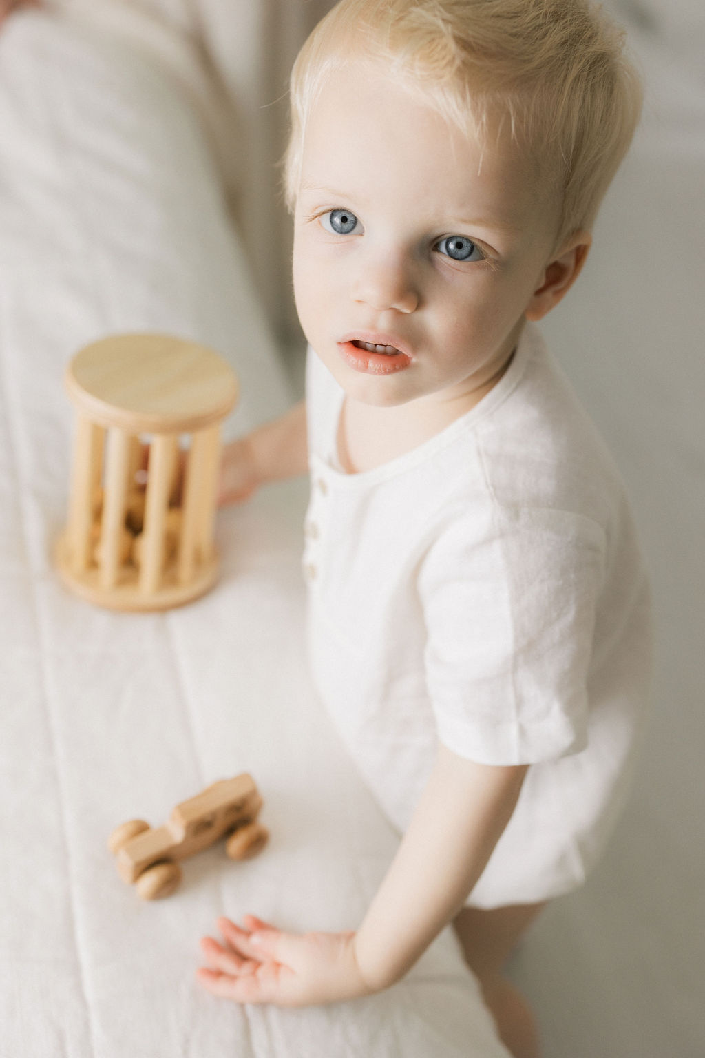 A blonde toddler boy plays on a bed with wooden toys in a white onesie after visiting toy stores nj