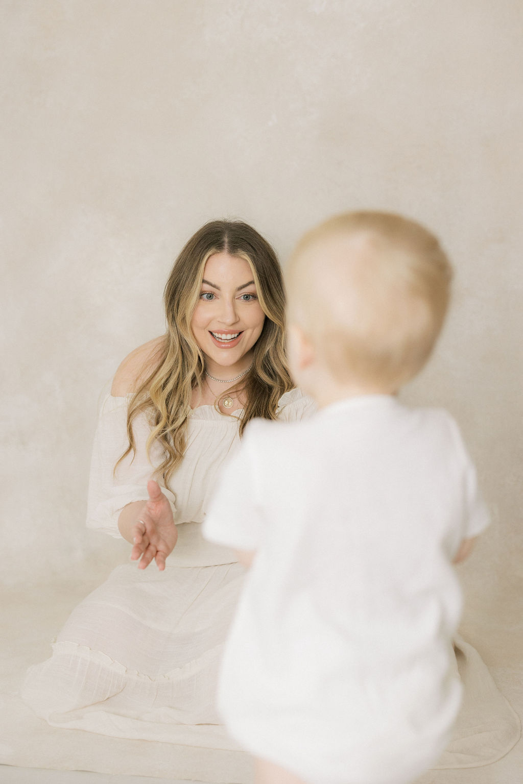 A happy mom in a white dress plays with her toddler son in a studio after using happy family after