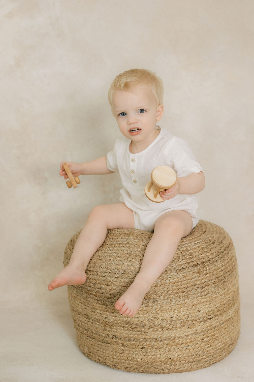 A toddler boy plays with wooden toys while sitting on a woven stool in a studio after meeting happy family after